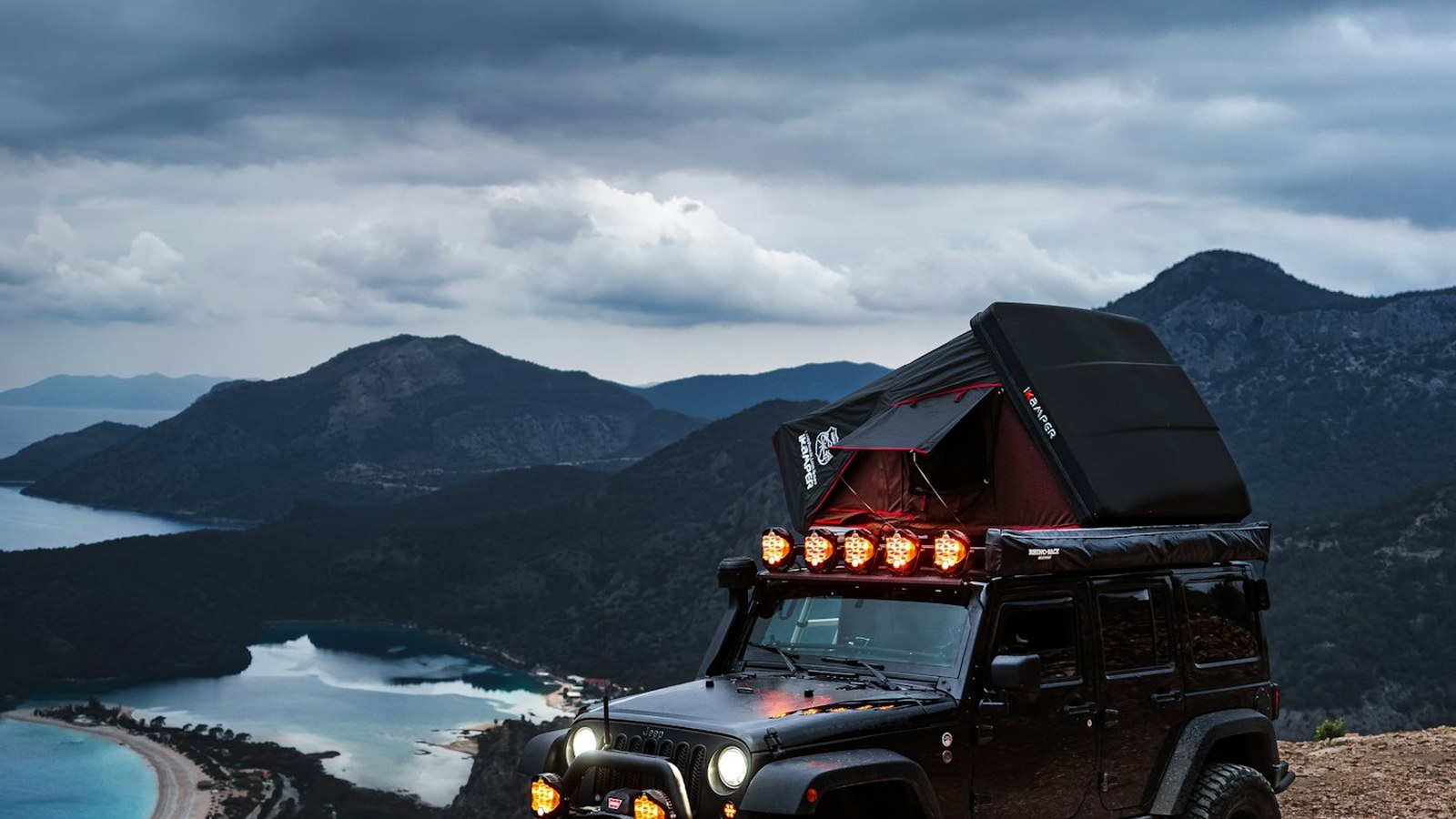 Jeep with a rooftop tent parked on a clifftop above a lagoon under a moody sky