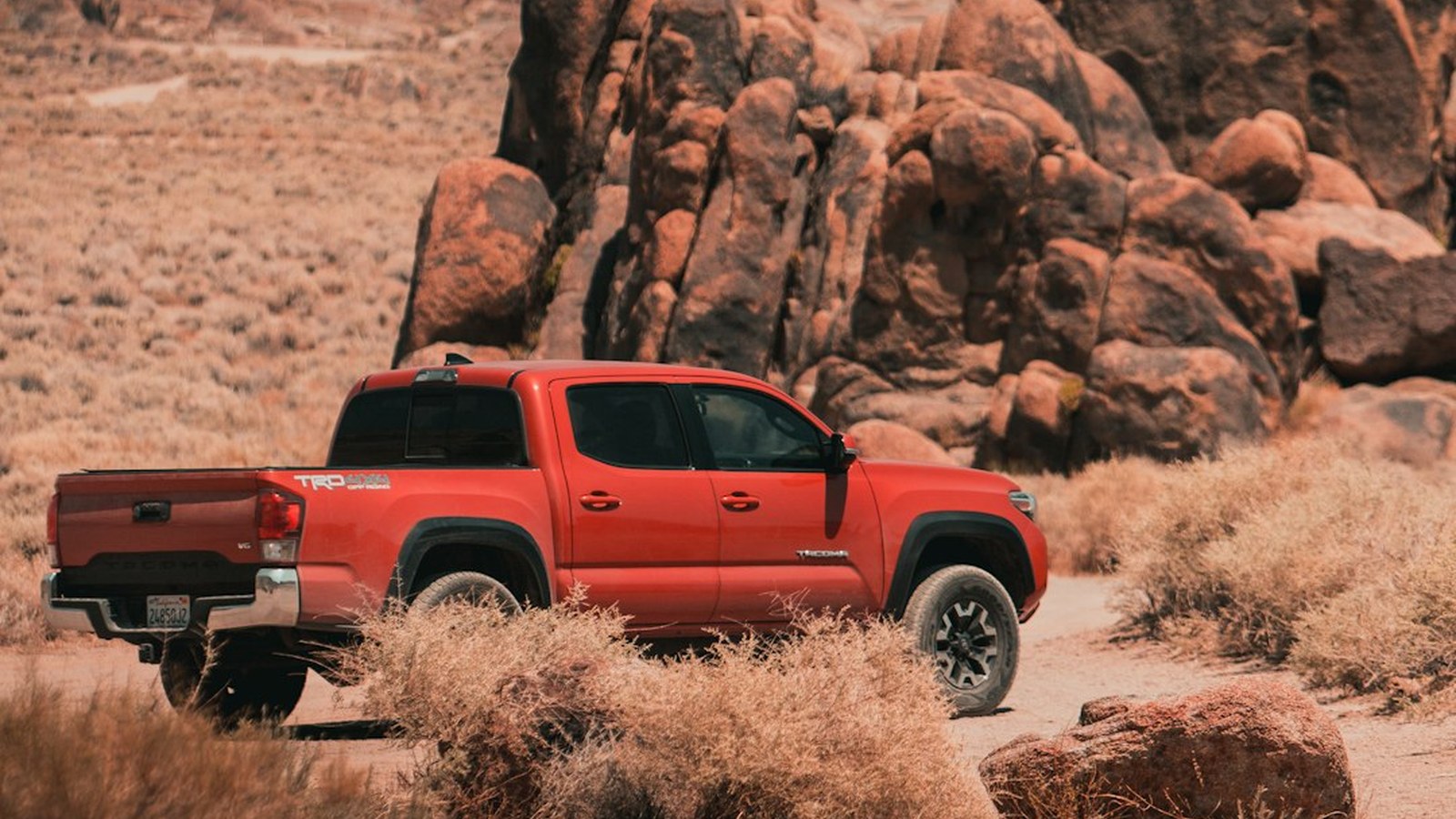 Red Toyota Tacoma on a sandy desert trail winding through golden brush and boulders