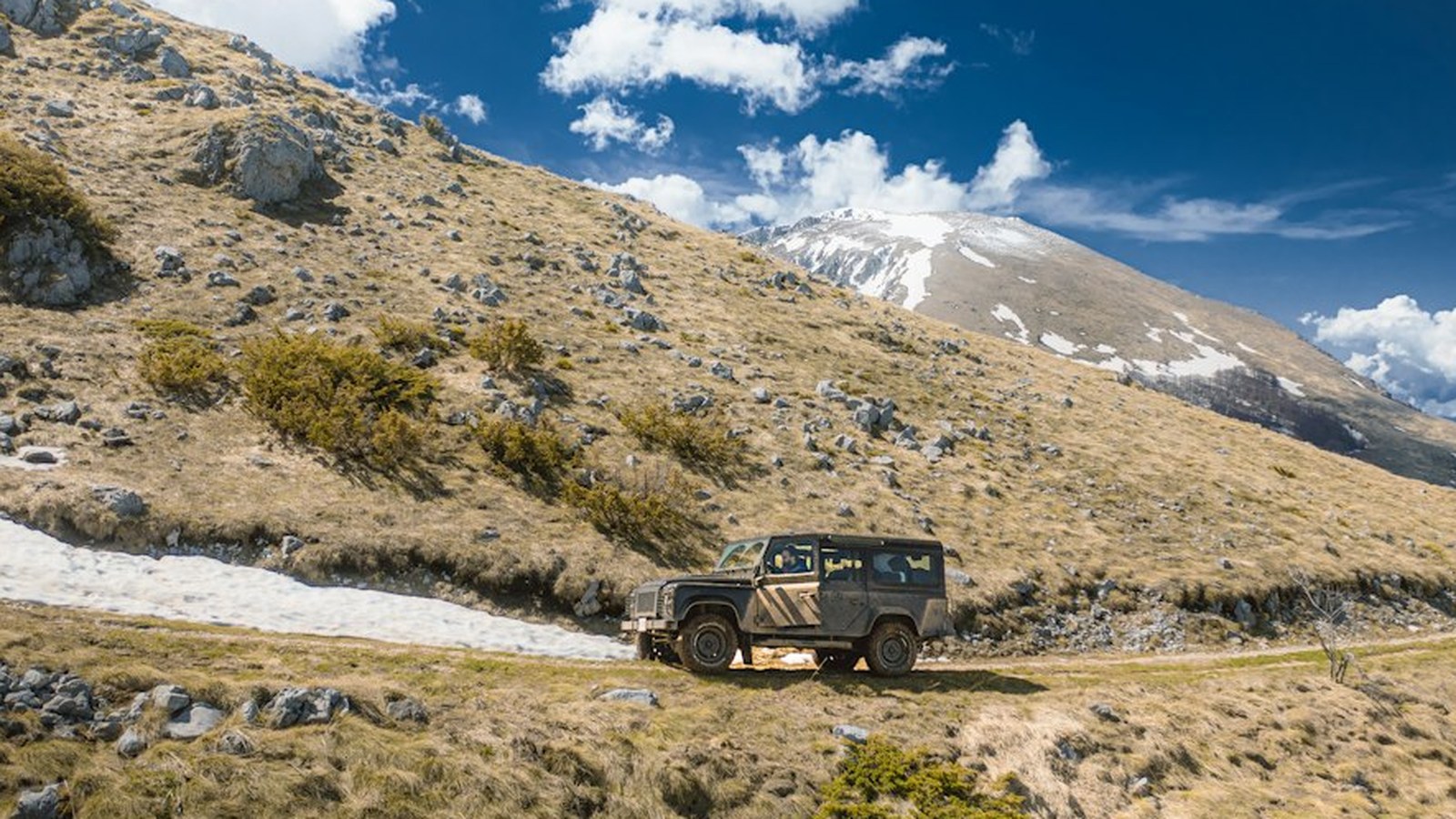 A Land Rover Defender on a rugged alpine dirt track with snow-dusted peaks behind it