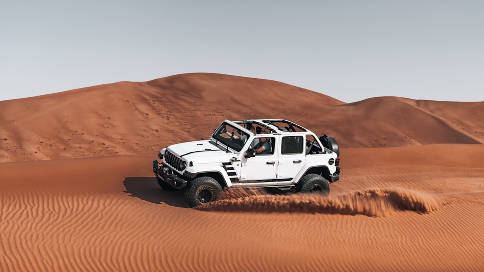 A white Jeep carving across a soft desert dune with sand spraying off the tires