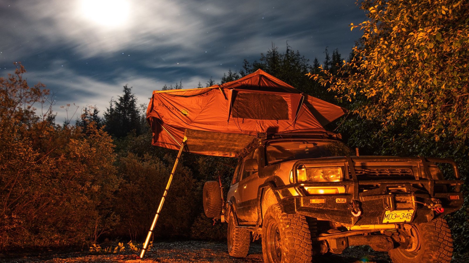 A 4x4 with a rooftop tent lit up at a nighttime overland campsite under a full moon