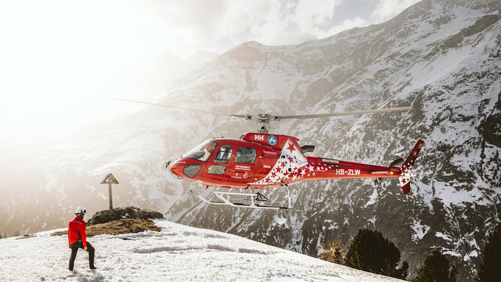 A red search and rescue helicopter approaching a person signaling in snowy mountain terrain