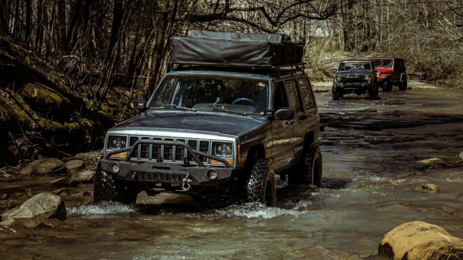 Lifted Jeep Cherokee XJ with a roof tent fording a rocky stream on a trail, convoy behind