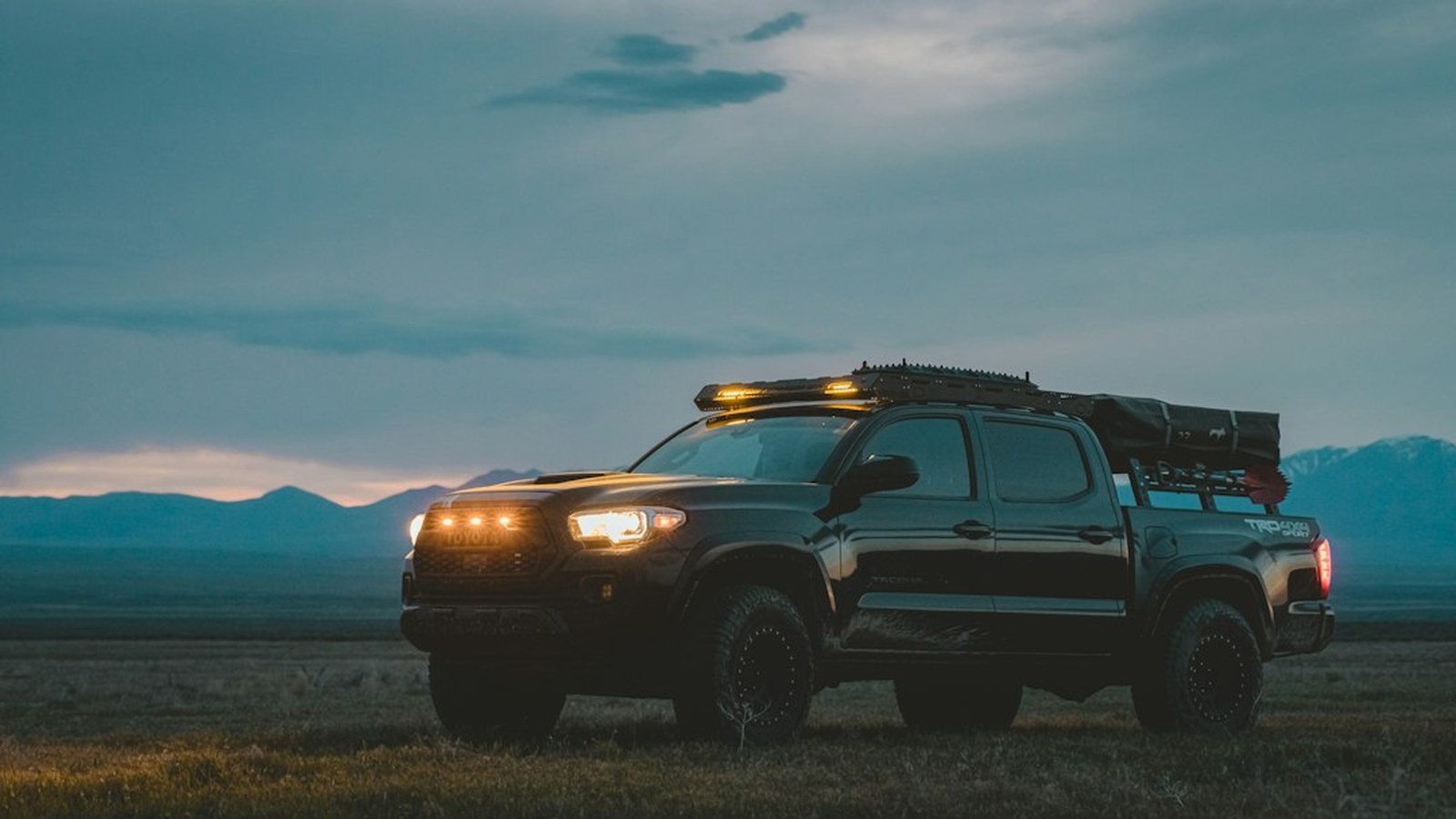 A black Toyota Tacoma overland build with roof rack and auxiliary lights on a grassy plain at dusk with distant mountains