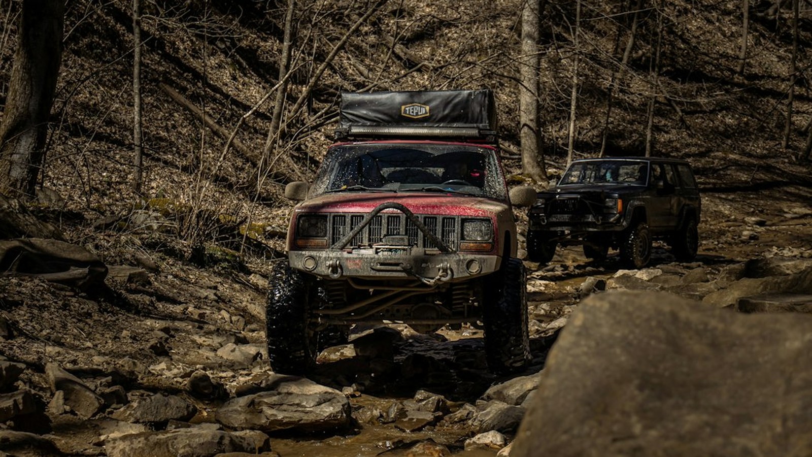 Two lifted Jeep Cherokee XJs crawling over a rocky creek bed through bare forest