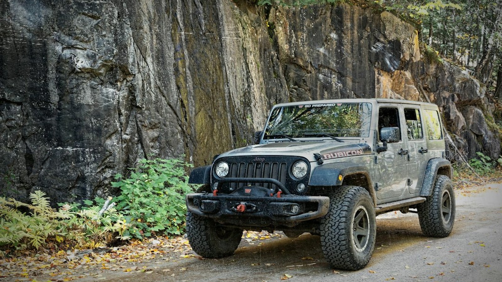 A gray Jeep Wrangler Rubicon with a winch bumper and off-road tires on a narrow rocky trail beside a stone wall
