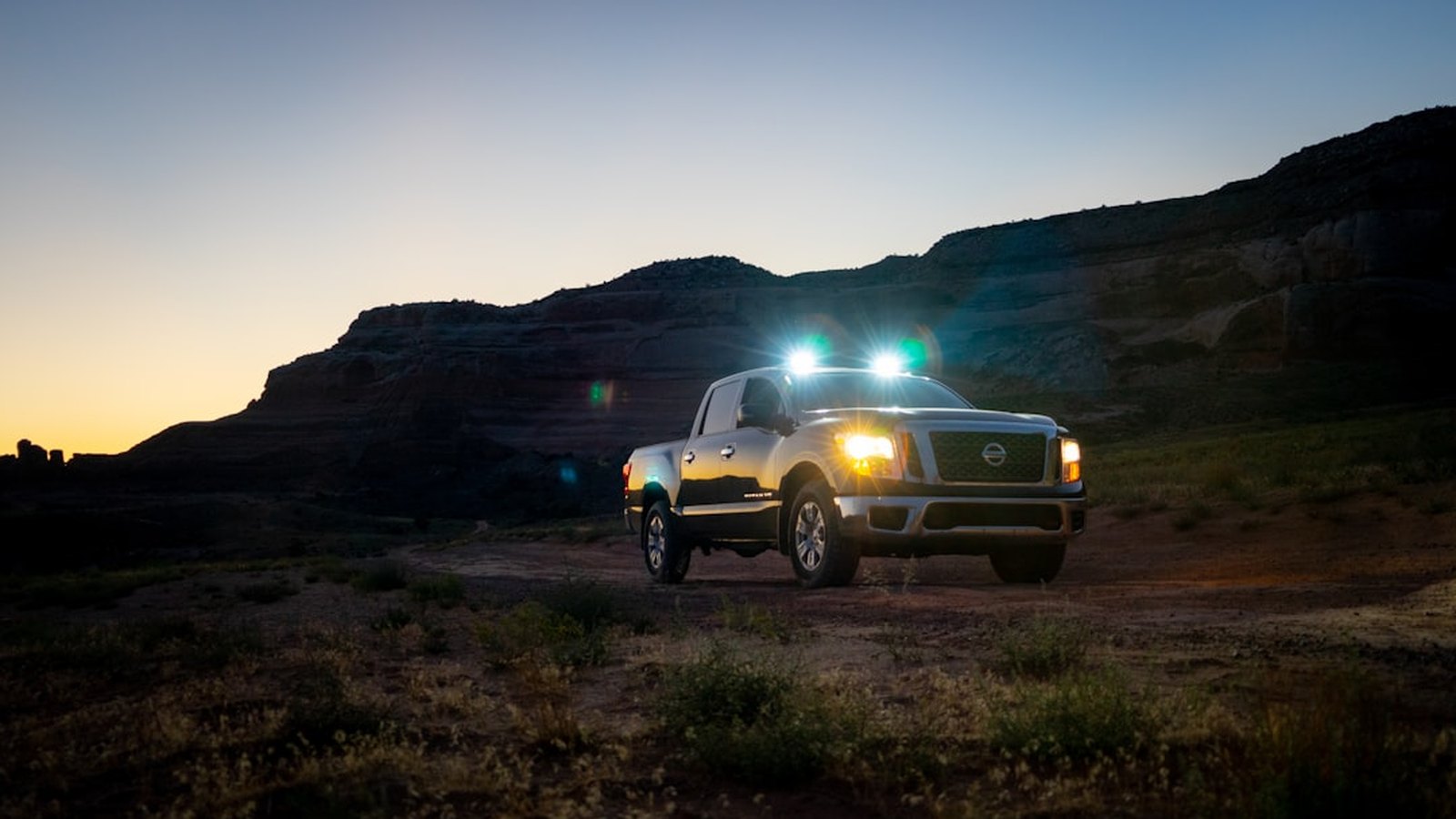 A 4x4 pickup at dusk with its roof lights and headlights glowing against a canyon backdrop