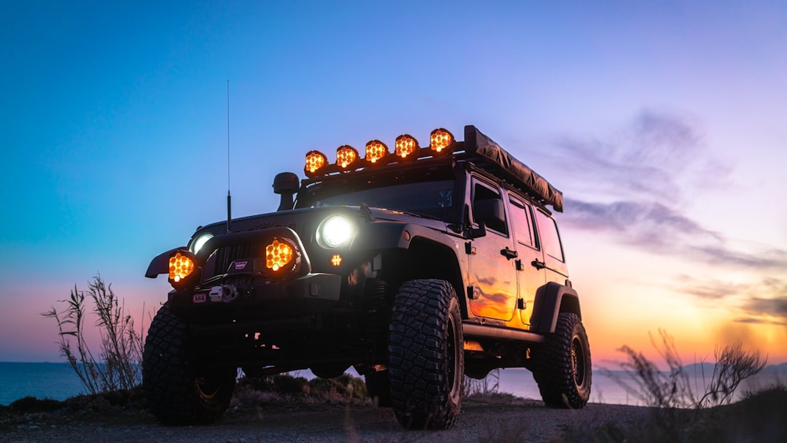 A lifted Jeep Wrangler on a coastal ridge at sunset with roof-mounted pod lights glowing