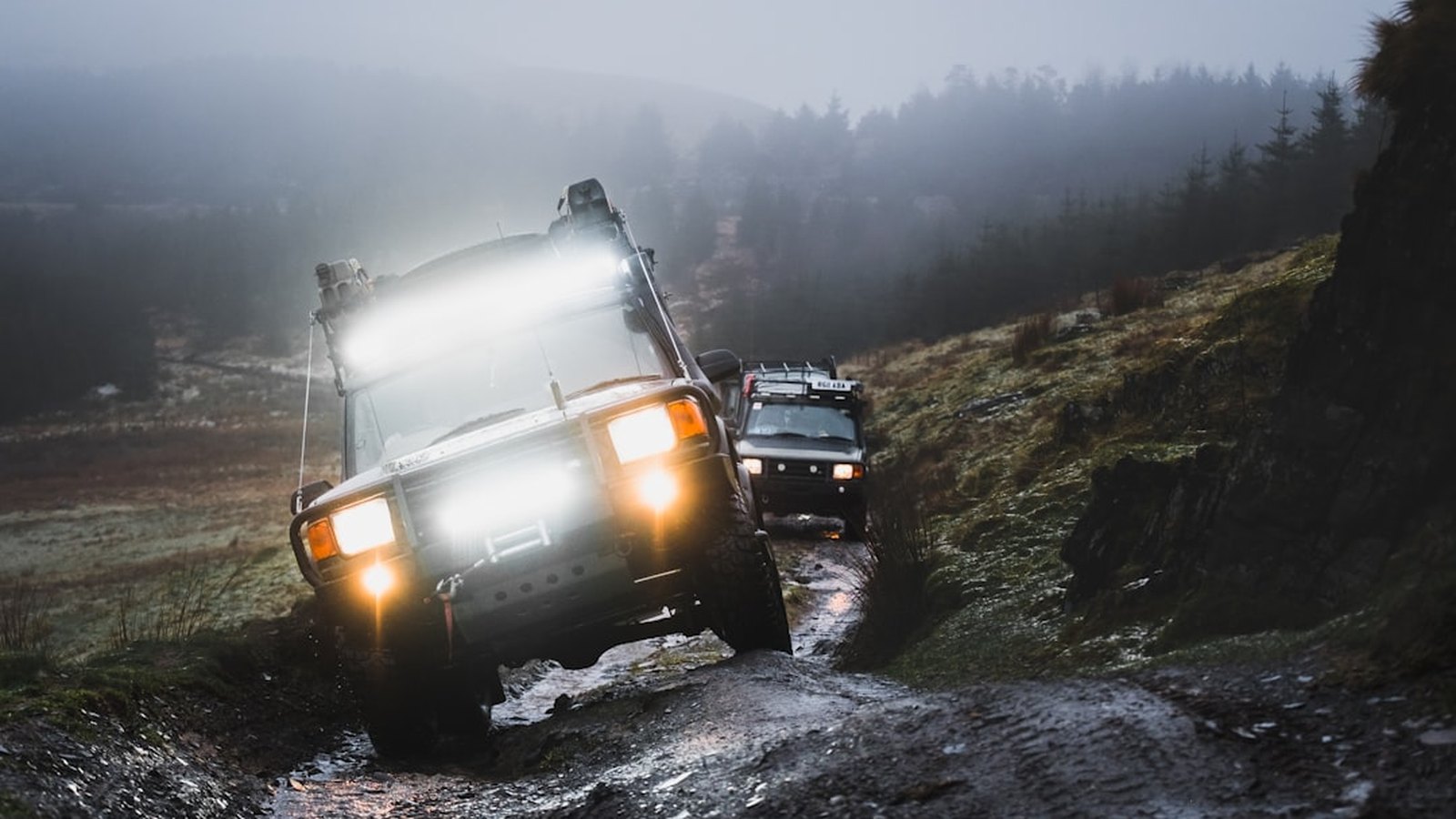 Two 4x4 rigs climbing a muddy off-camber trail at dusk with light bars and spot lights firing through the haze