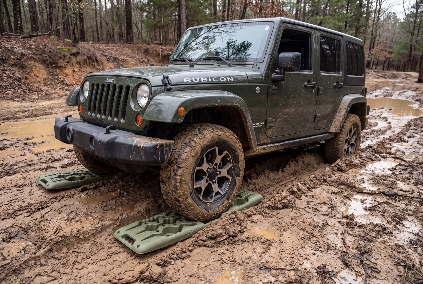 A muddy Jeep Wrangler bogged down in deep ruts with traction boards set under the front tire