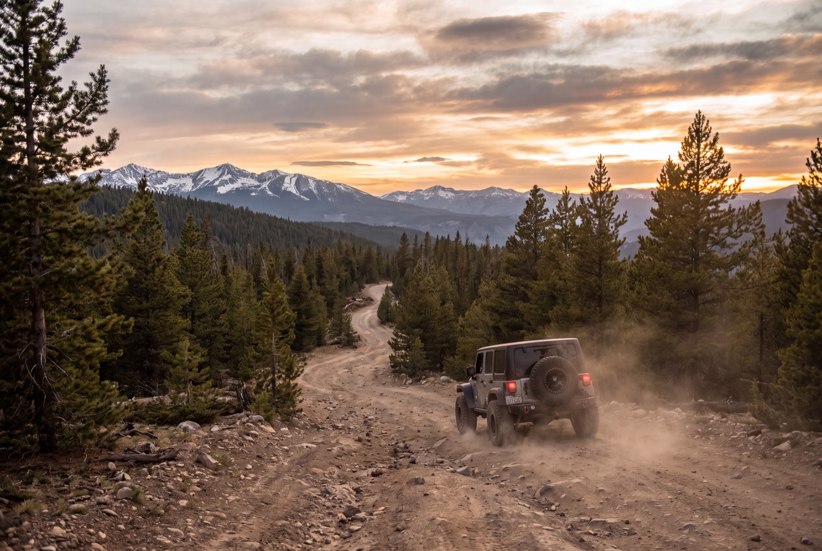 A Jeep Wrangler driving away down a dusty mountain road at sunset