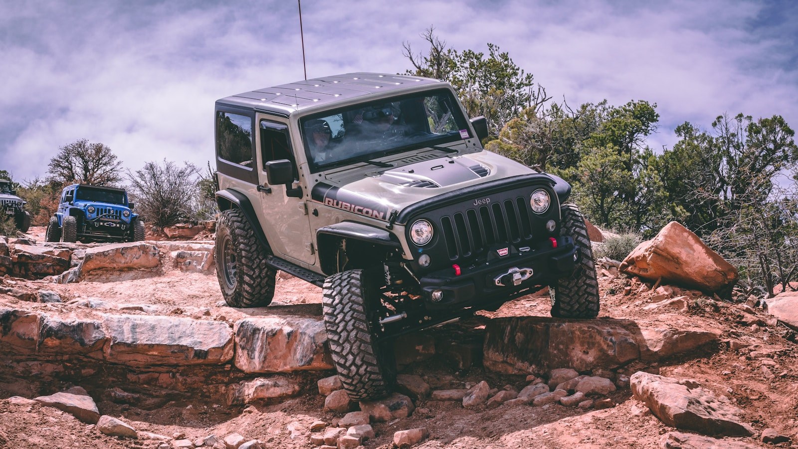 A tan Jeep Wrangler Rubicon flexing up onto large red boulders on a rock trail