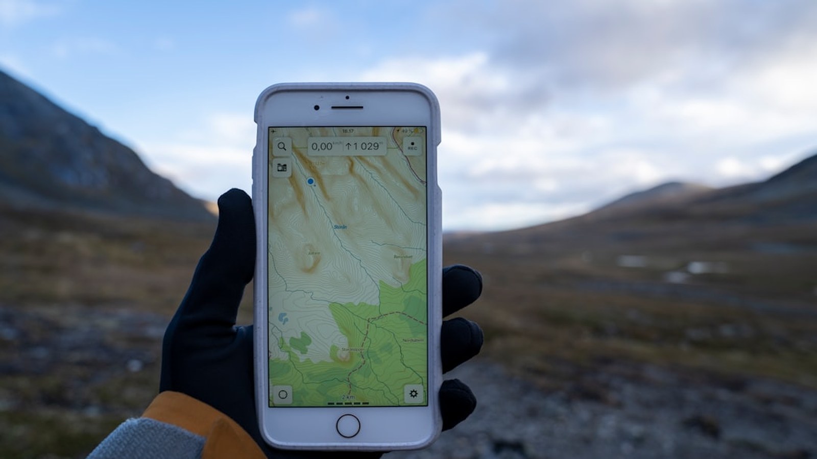 A gloved hand holding a phone running a GPS navigation app on a mountain trail