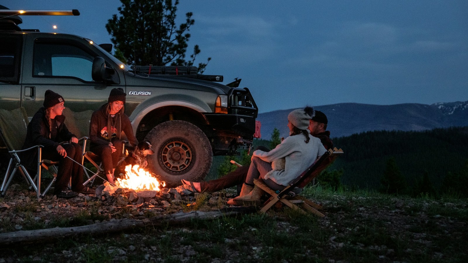 Overlanders gathered around a campfire beside a rig at night with camp lights glowing