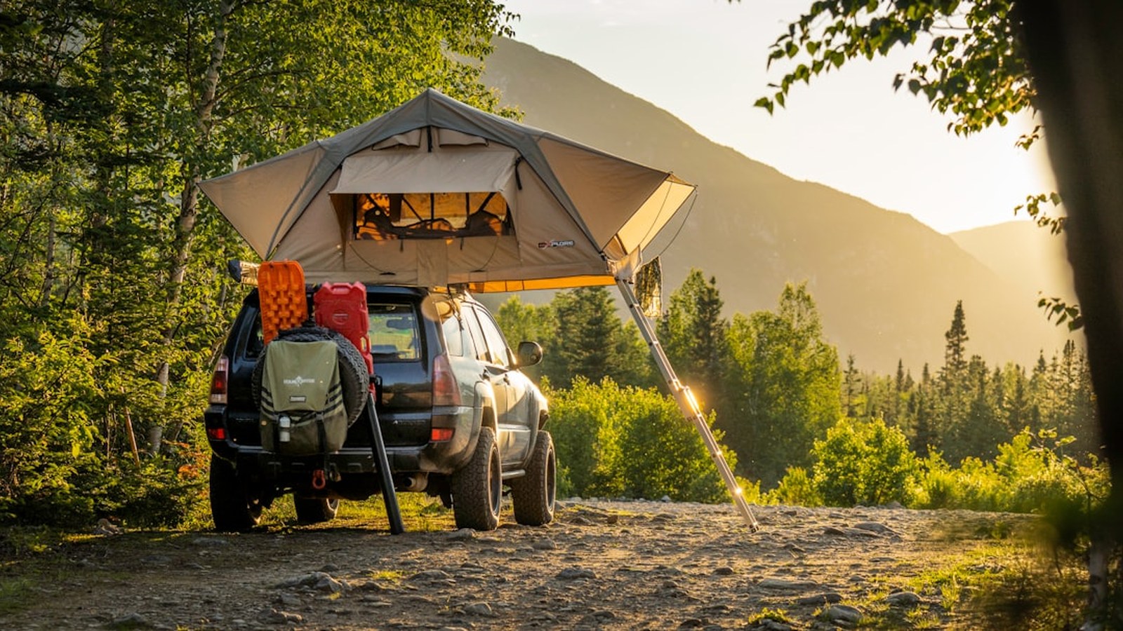 A loaded overland 4x4 with a deployed rooftop tent, traction boards, and fuel cans set up in remote backcountry at golden hour.