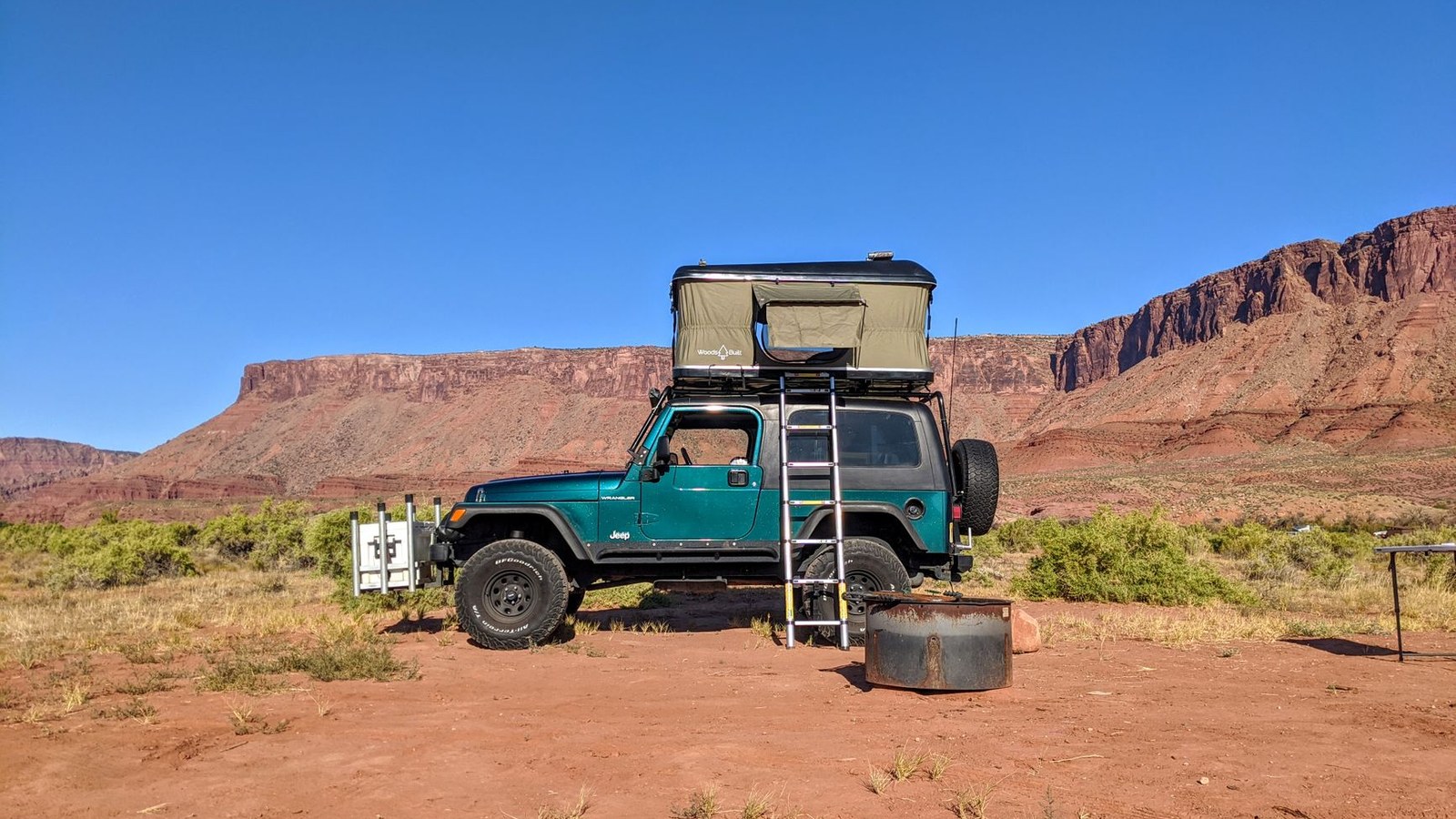 A Jeep Wrangler with a deployed rooftop tent parked in red-rock canyon country on a family overland camp.