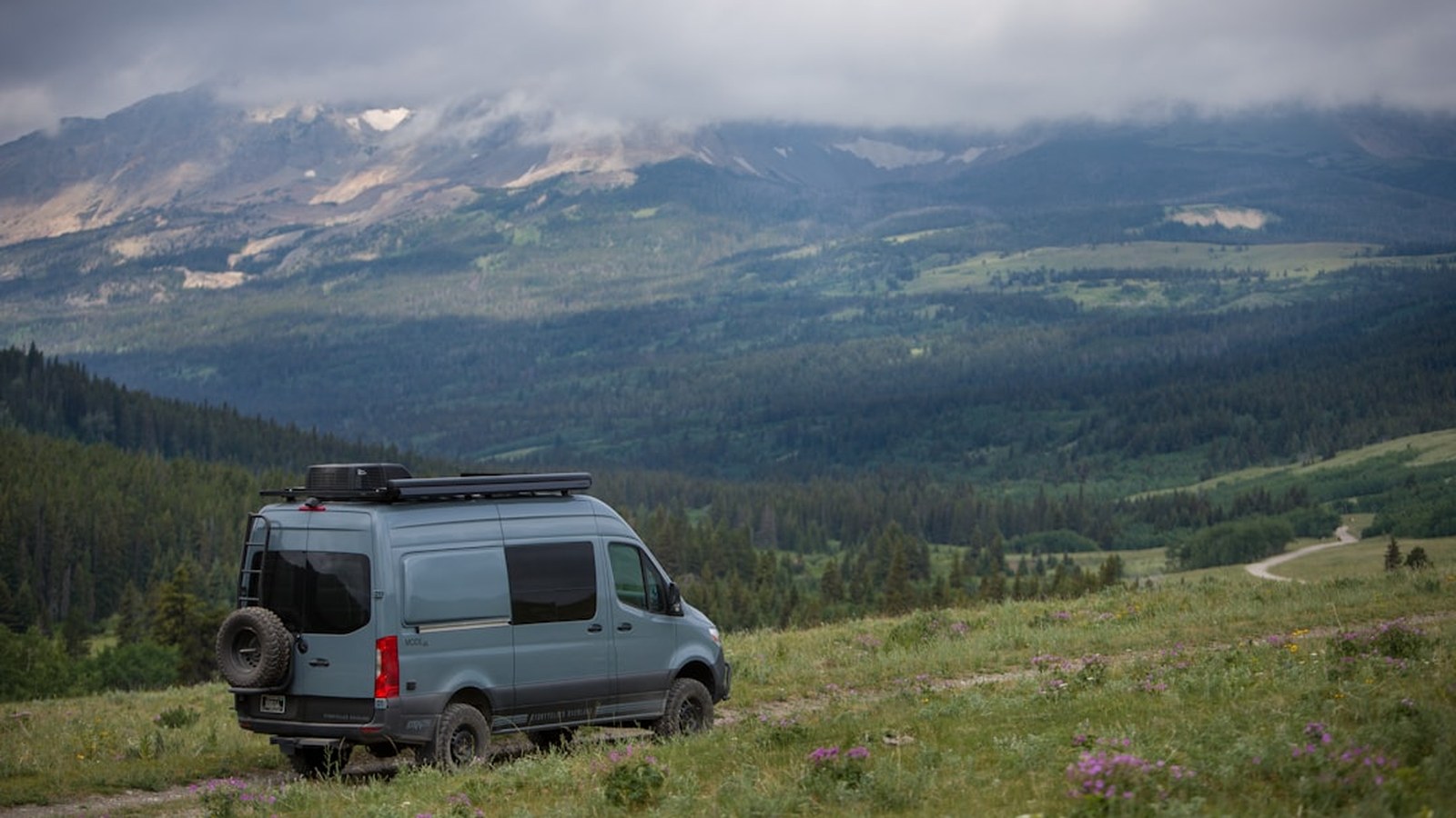 Overland rig parked in an alpine meadow beneath dramatic cloud-covered peaks