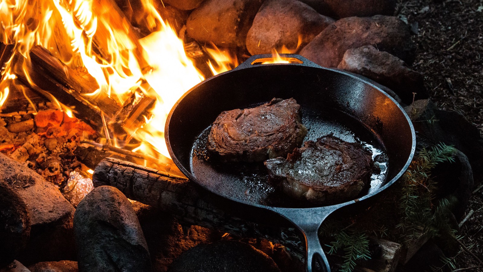 Steaks searing in a cast-iron skillet at the edge of a live campfire