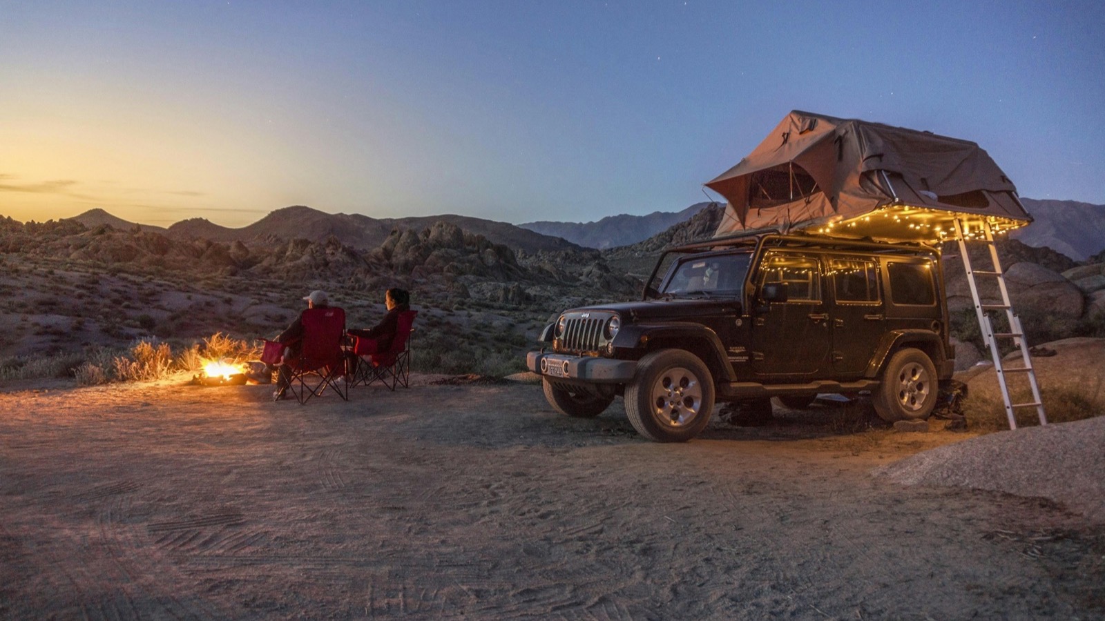 A Jeep Wrangler with a rooftop tent at a wild desert camp among granite boulders at dusk