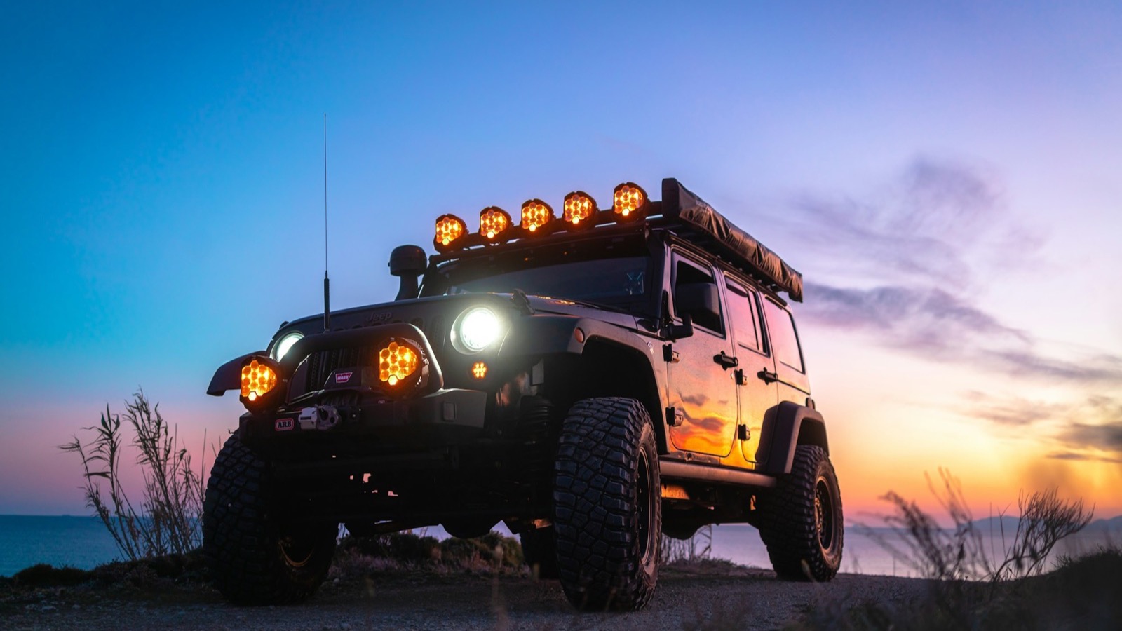 A kitted out Jeep parked at a trailhead at dusk with its lights on