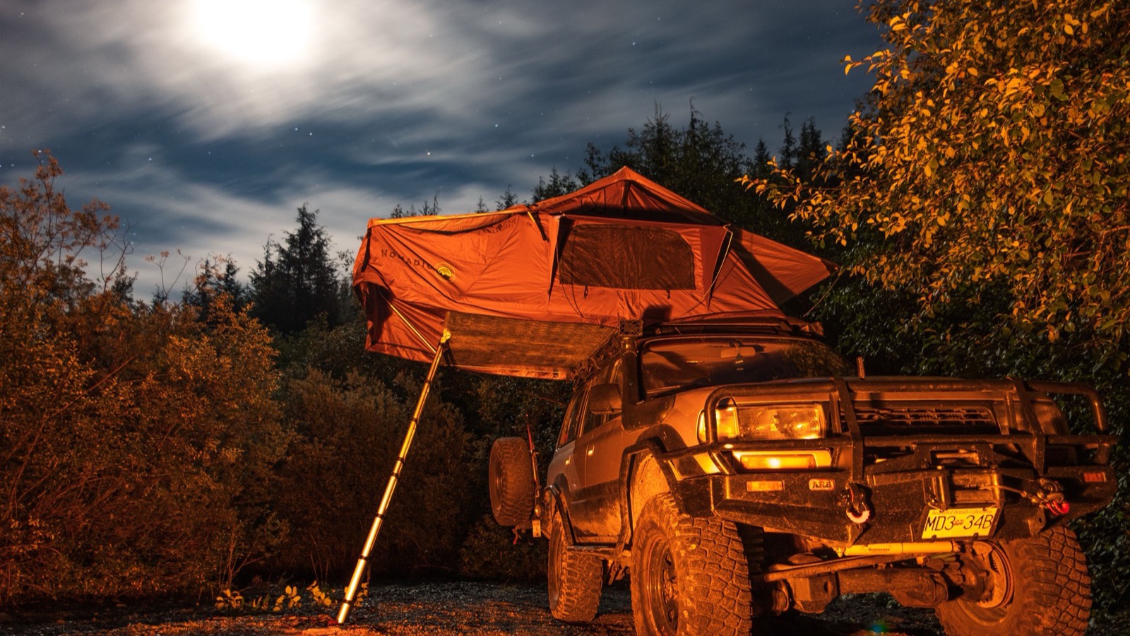 A 4x4 with an orange rooftop tent deployed under a moonlit sky with a campfire glow