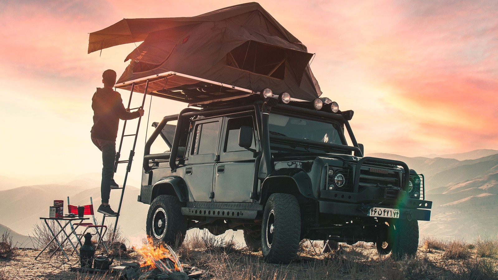 A Land Rover Defender with a rooftop tent deployed and a campfire below pink sunset peaks