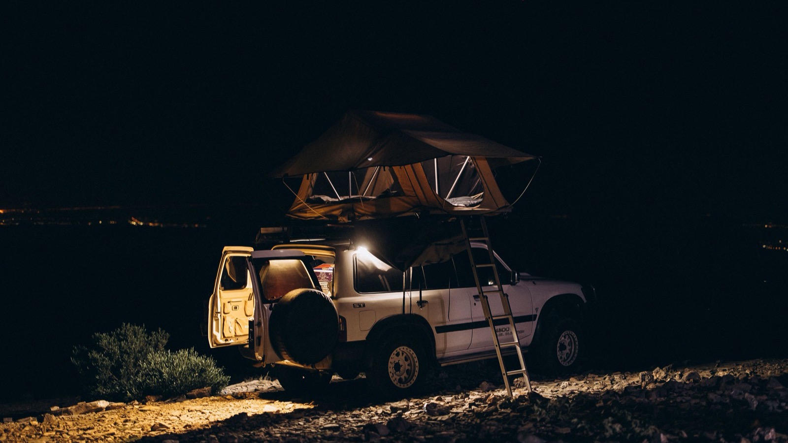 A 4x4 with a rooftop tent deployed and ladder down at a night camp overlooking distant lights