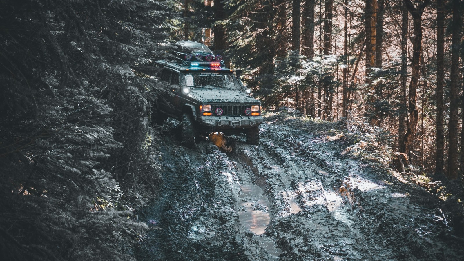 A muddy 4x4 crawling a wet rutted trail with headlights cutting the gloom
