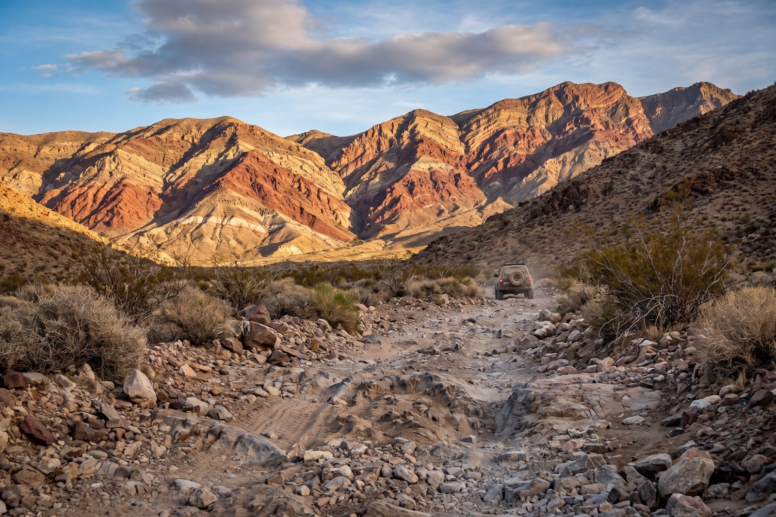 A rugged sandy 4x4 two-track climbing through the folded, banded red and tan rock of the Panamint Range in Death Valley, a lone pickup driving toward the mountains in warm afternoon light