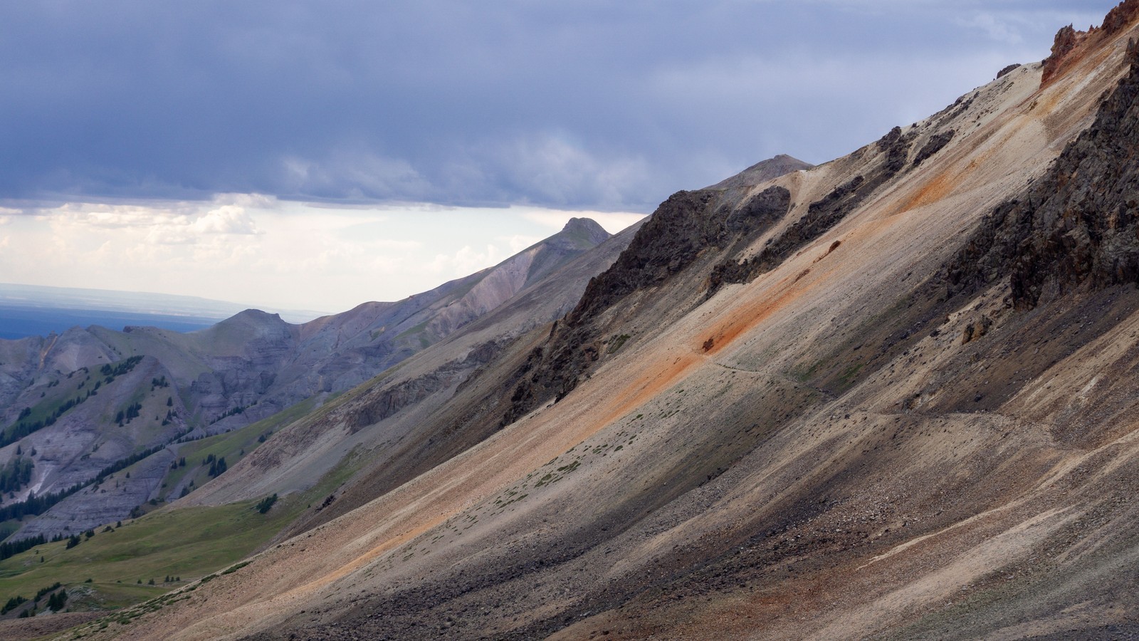 A switchback trail cut across a mineral-stained scree slope under a brooding sky high above treeline near Cinnamon Pass