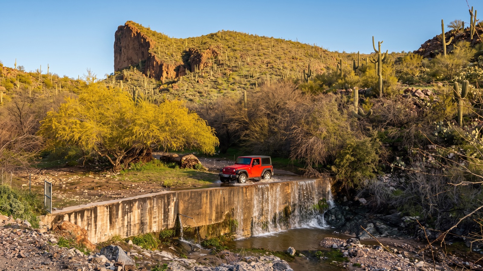 A shallow creek flowing over a concrete low-water crossing, a saguaro-studded desert butte rising behind it.