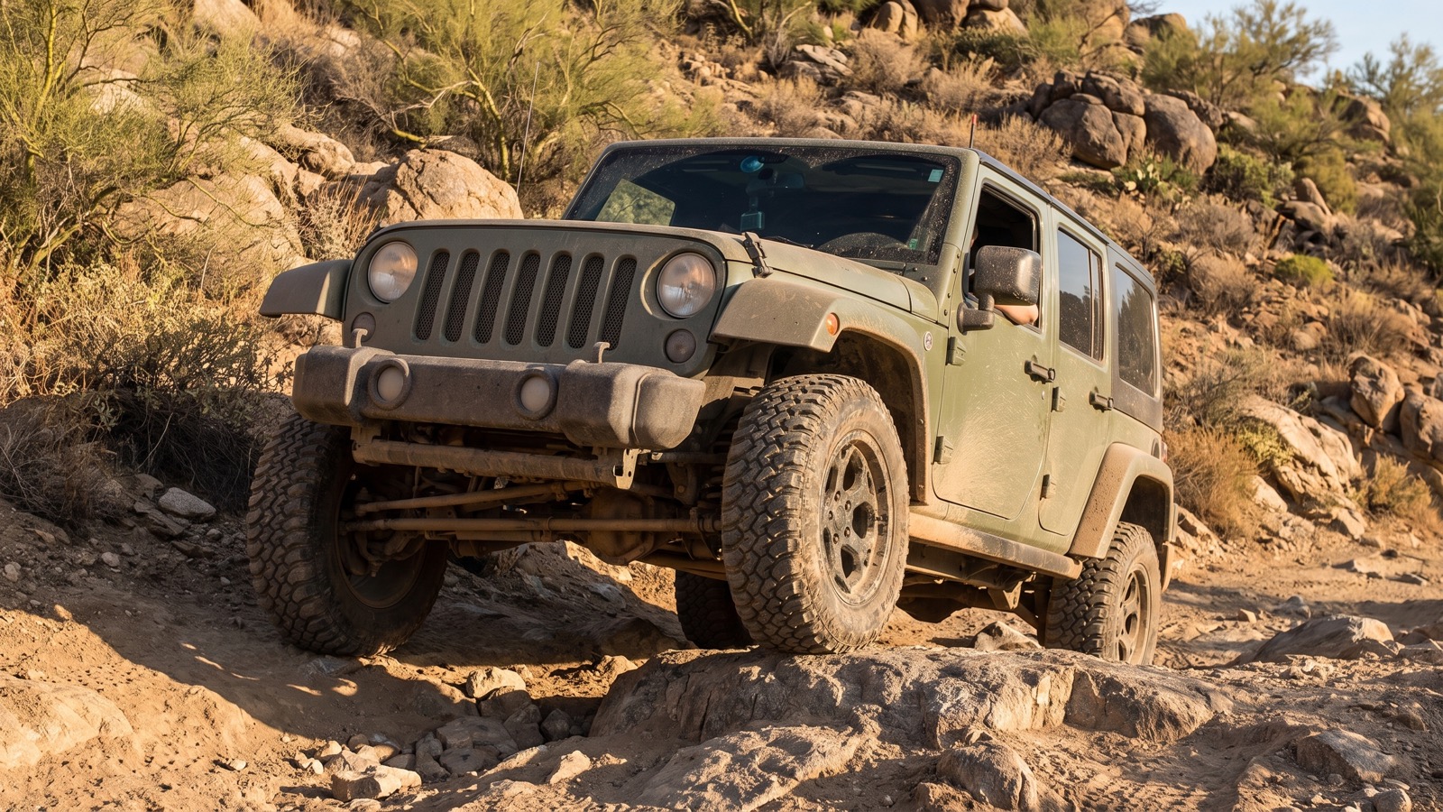 A green Jeep Wrangler flexing over a rocky ledge obstacle on the rugged climb, boulders and dry brush all around.