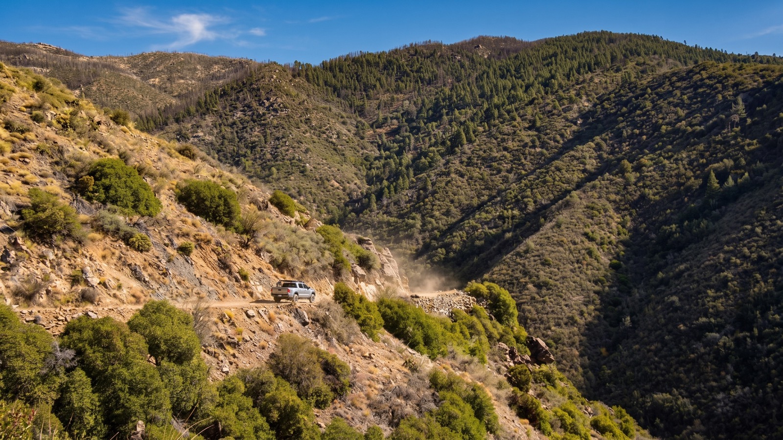 A narrow rocky shelf road cut into a steep hillside above a deep forested canyon, a rig raising dust as it climbs.