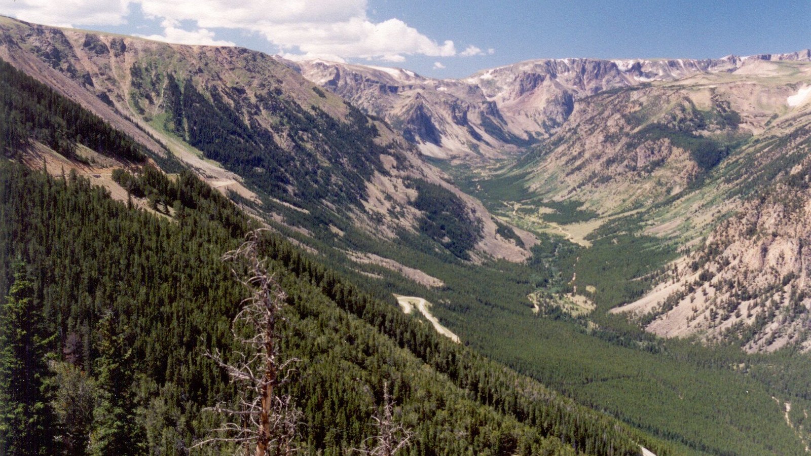 A deep U-shaped glacial valley in the Beartooth Mountains, with the highway threading the forested valley floor far below the rugged peaks