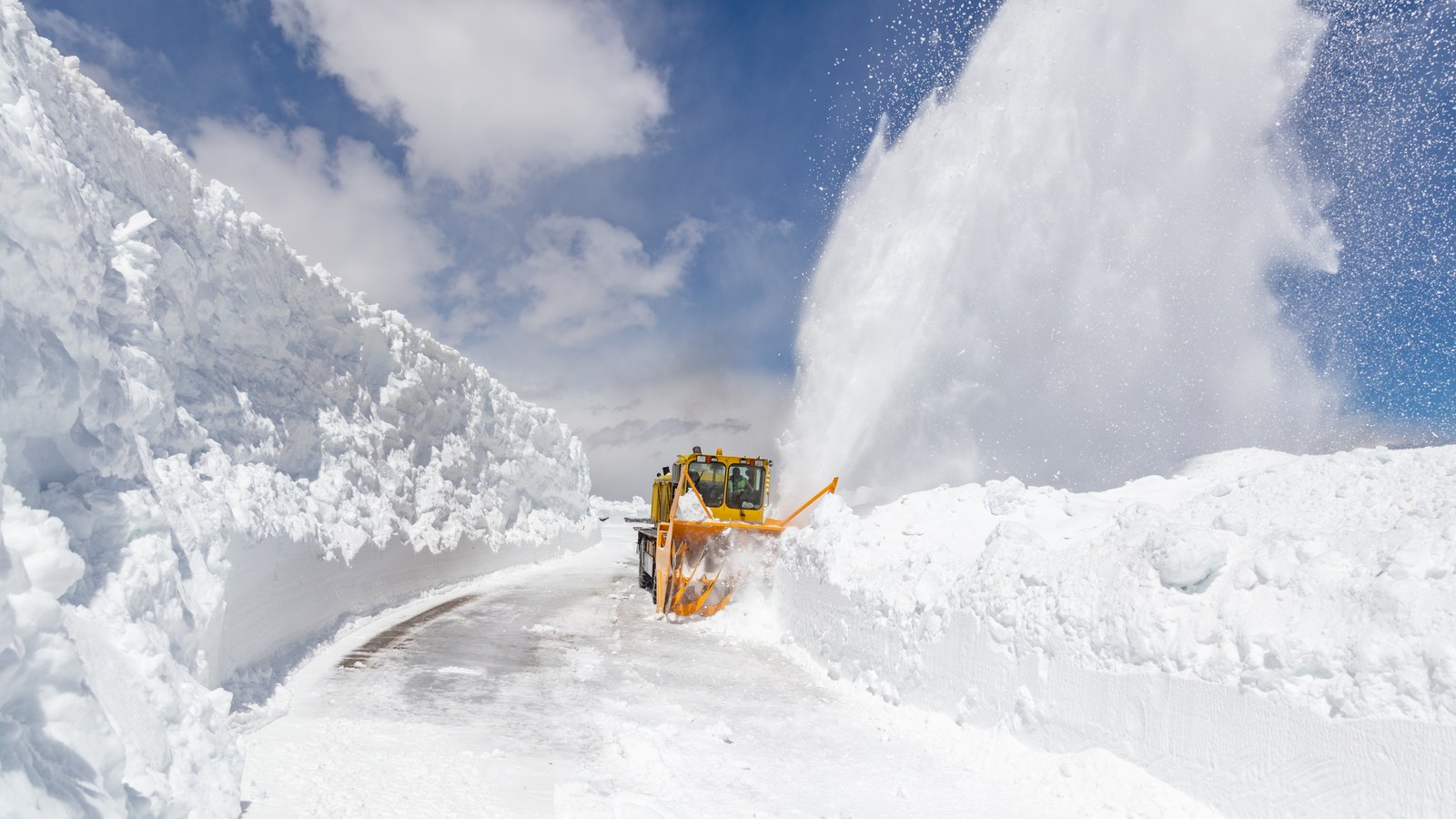 A rotary snowplow blasting through a corridor of towering snow walls to open the Beartooth Highway in spring, with banks taller than the road