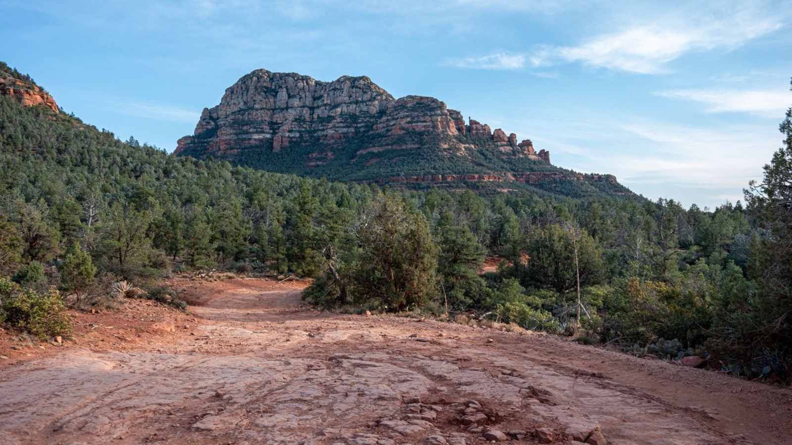 Red slickrock and a dirt off-road track winding toward a layered Sedona butte under a blue sky