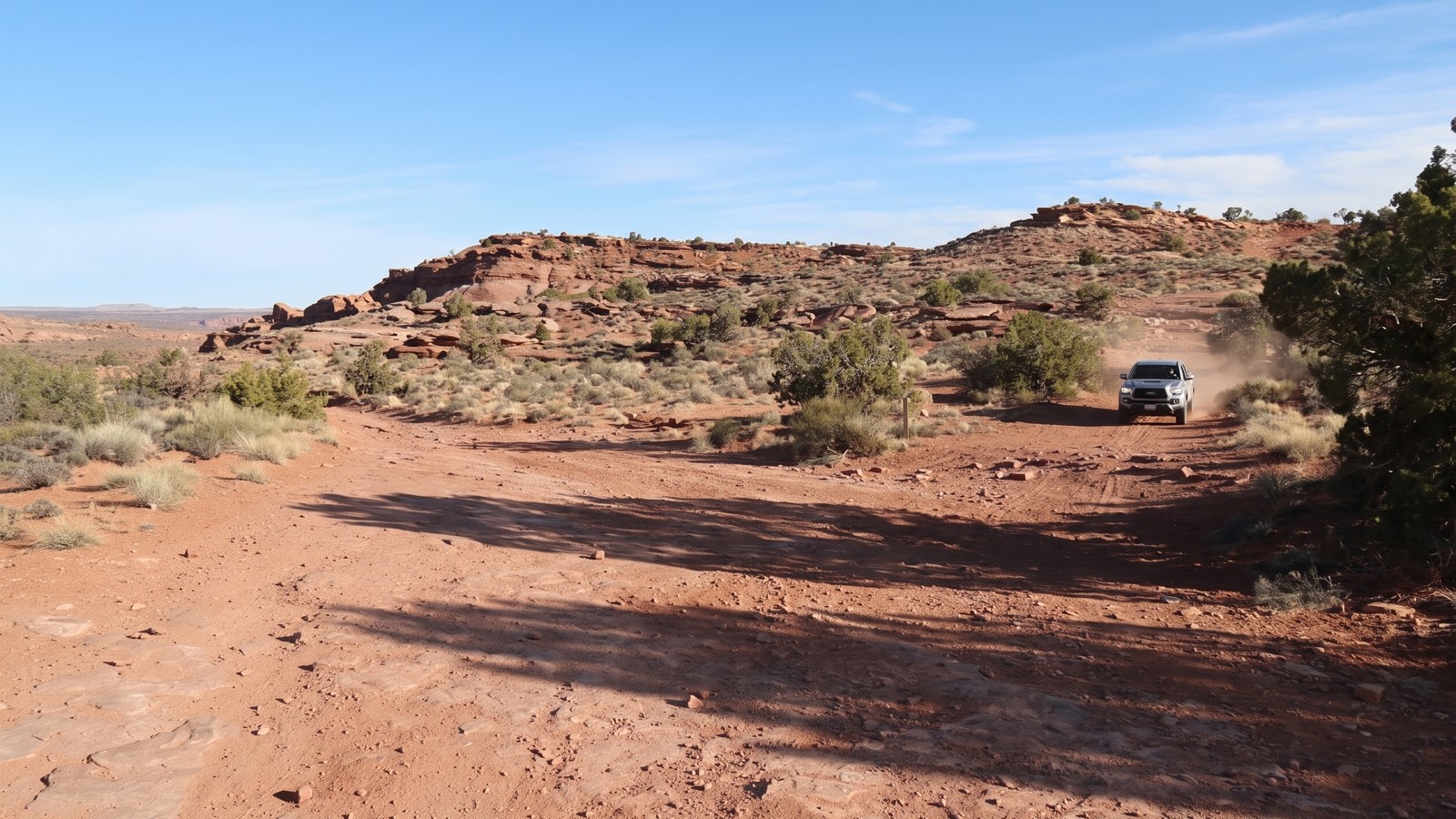 A silver Toyota Tacoma navigates the open mesa trail on Amasa Back with juniper trees and distant canyon rims