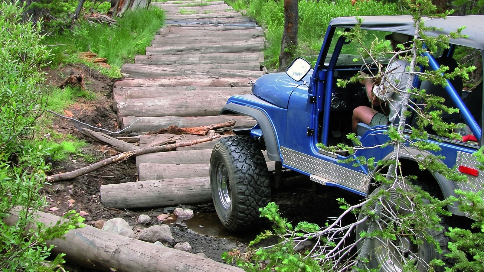 A short-wheelbase Jeep crossing a log corduroy bridge over a creek in the tight forest of the Dusy-Ershim Trail