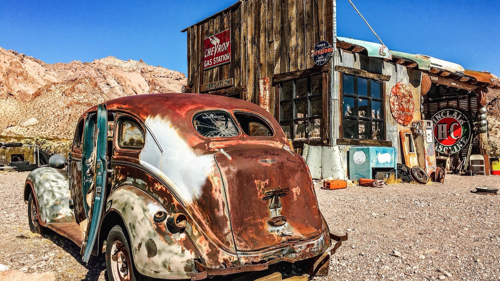 A rusted vintage car parked in front of a weathered Chevron gas-station facade with classic enamel signs at the Nelson ghost town in El Dorado Canyon, Nevada