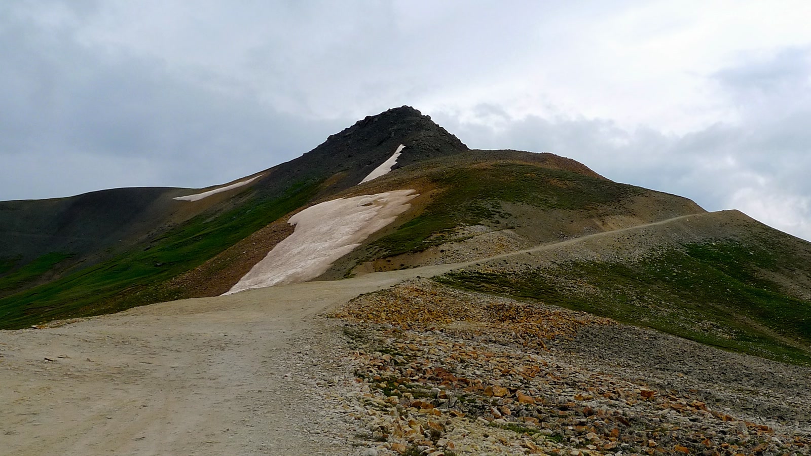 The Engineer Pass shelf road climbing a bare high-alpine ridge toward the rocky summit, snow lingering in the shadows