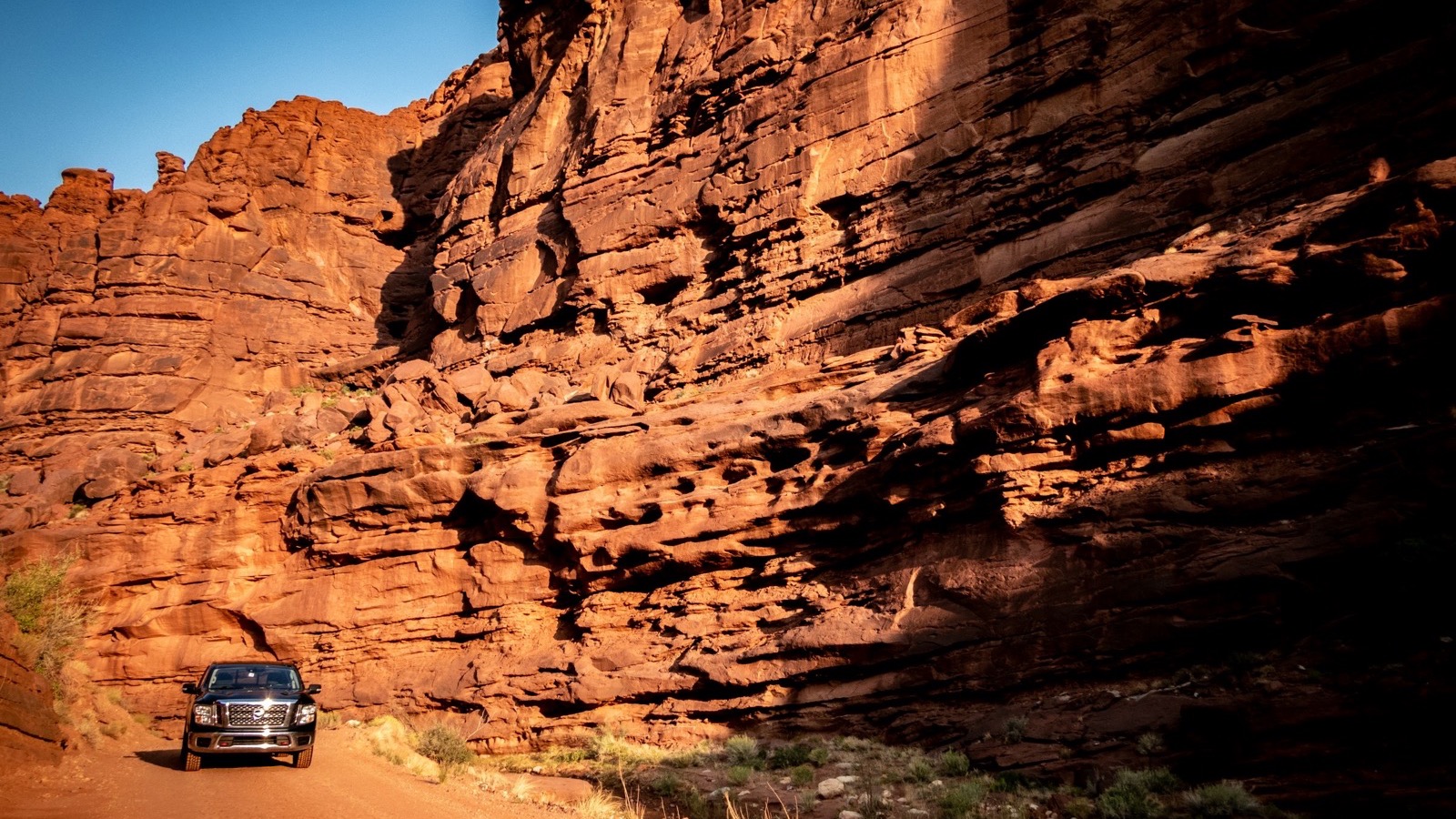 A 4x4 on a red dirt road hugging towering Moab sandstone walls in warm light