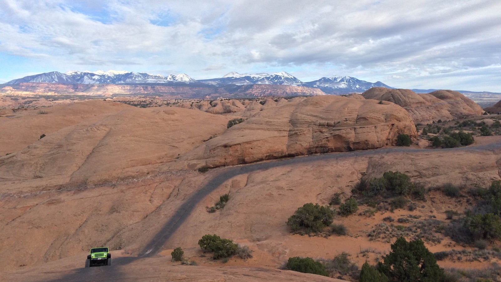 A green Jeep Wrangler on Moab slickrock with the snow capped La Sal Mountains behind it