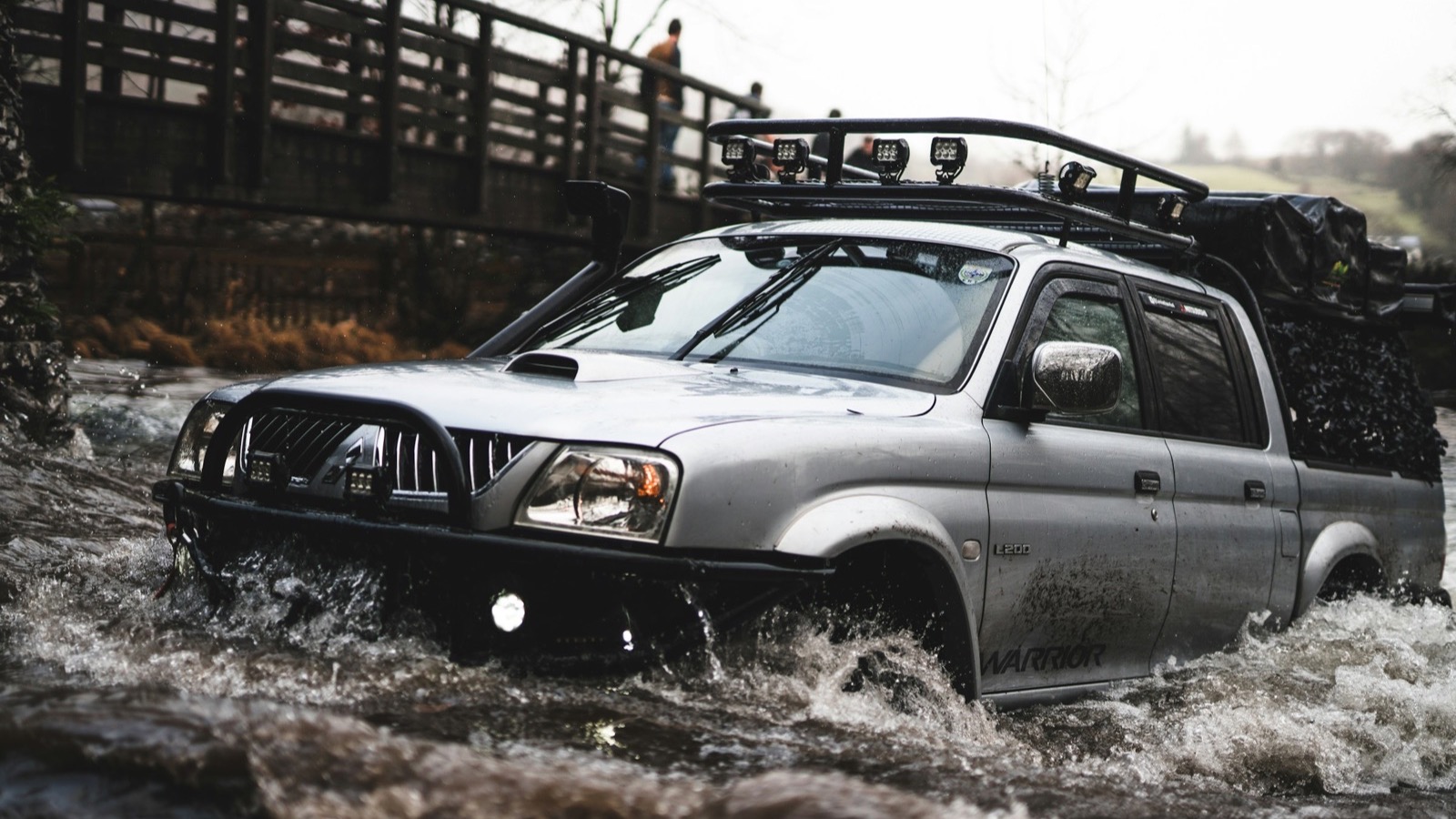 A kitted 4x4 powering through a deep river crossing with water spraying off the bumper, the kind of crossing Fordyce demands