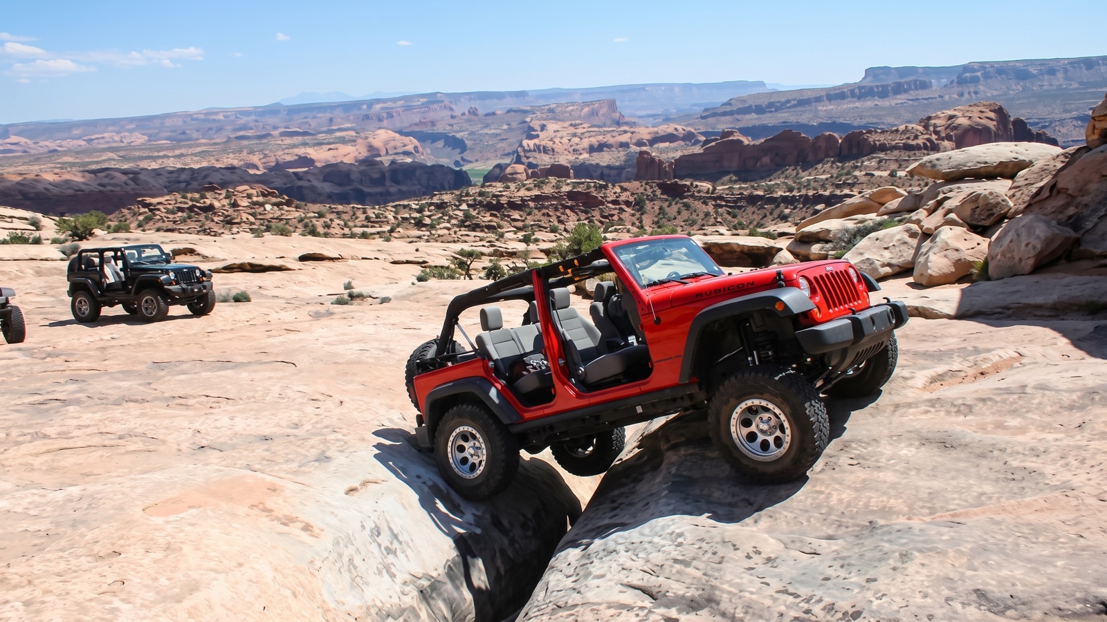 Two modified Jeep Wranglers crawling tan Moab slickrock on Golden Spike Trail with a sweeping canyon and mesa panorama behind