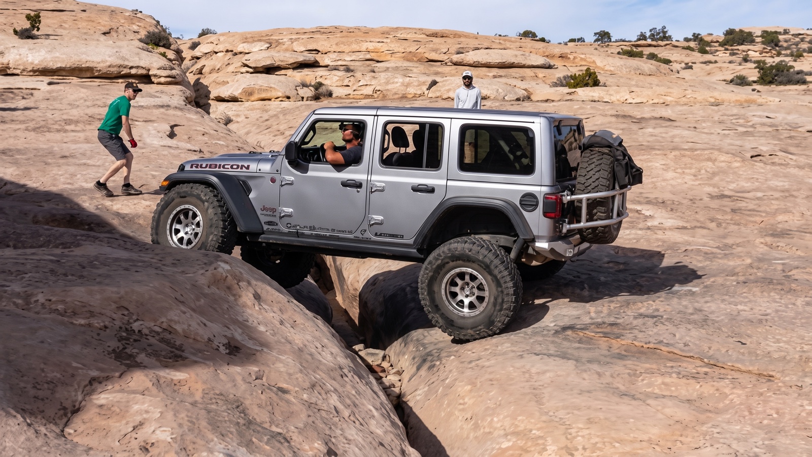 A silver Jeep Wrangler Rubicon dropping a wheel into the Golden Crack while a spotter guides the line on Golden Spike Trail
