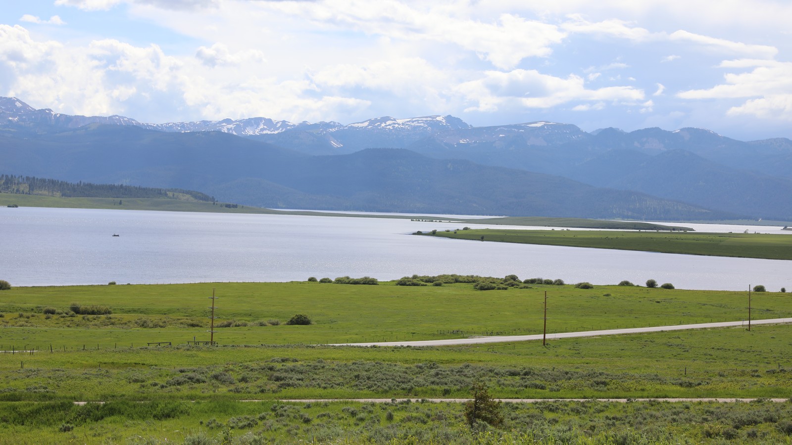 Wide view across Hebgen Lake with a shoreline gravel road, green meadow, and snow-streaked Montana mountains behind