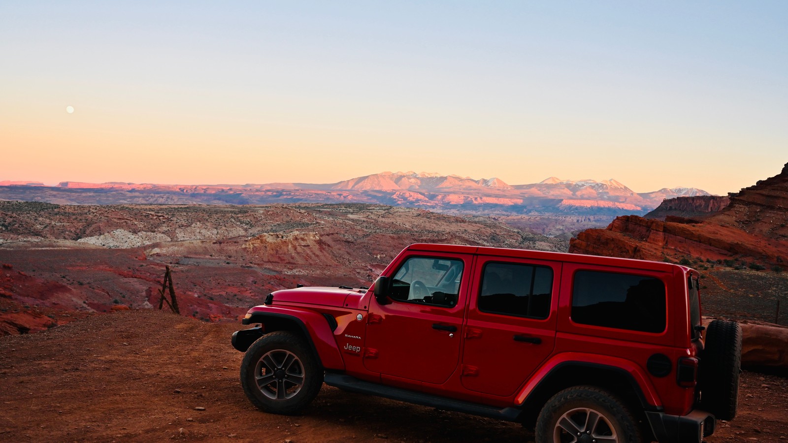 A red Jeep Wrangler on red rock at dawn with snow-dusted desert peaks and the moon on the horizon