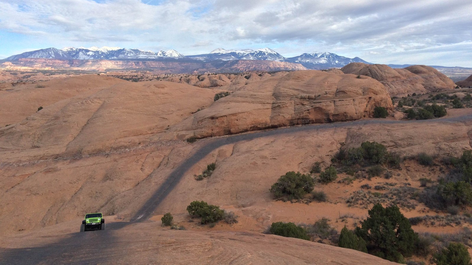 A green Jeep on the painted slickrock route of Hell's Revenge with the snow-capped La Sal Mountains across the horizon