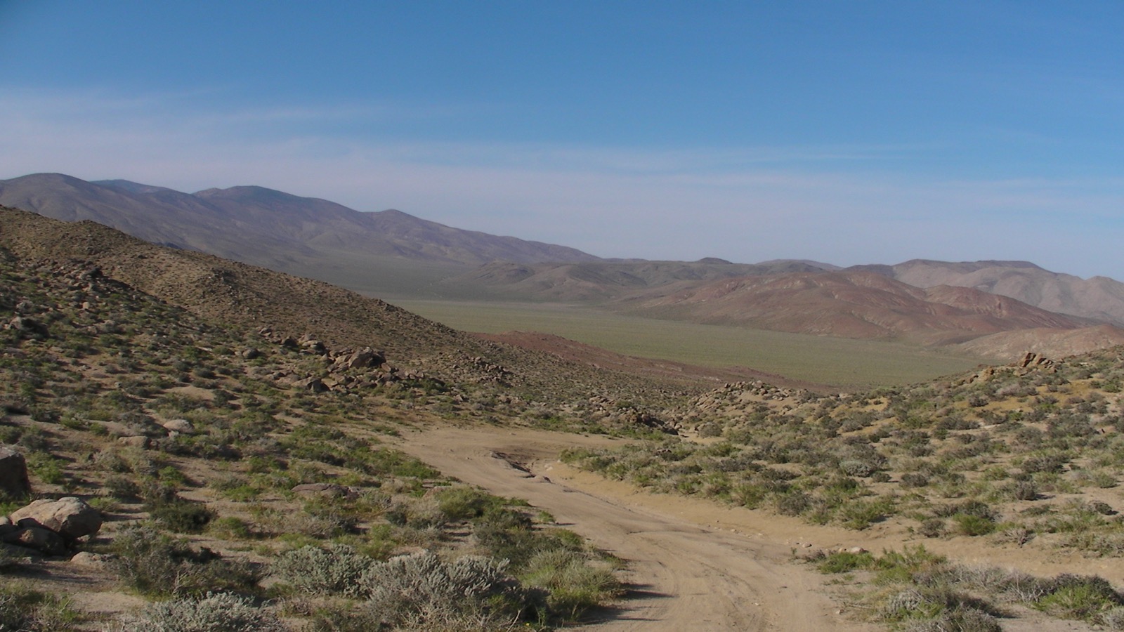 An open desert wash in the Panamint Range with a rocky two-track trail winding through scattered boulders and low scrub toward distant mountains under a clear sky