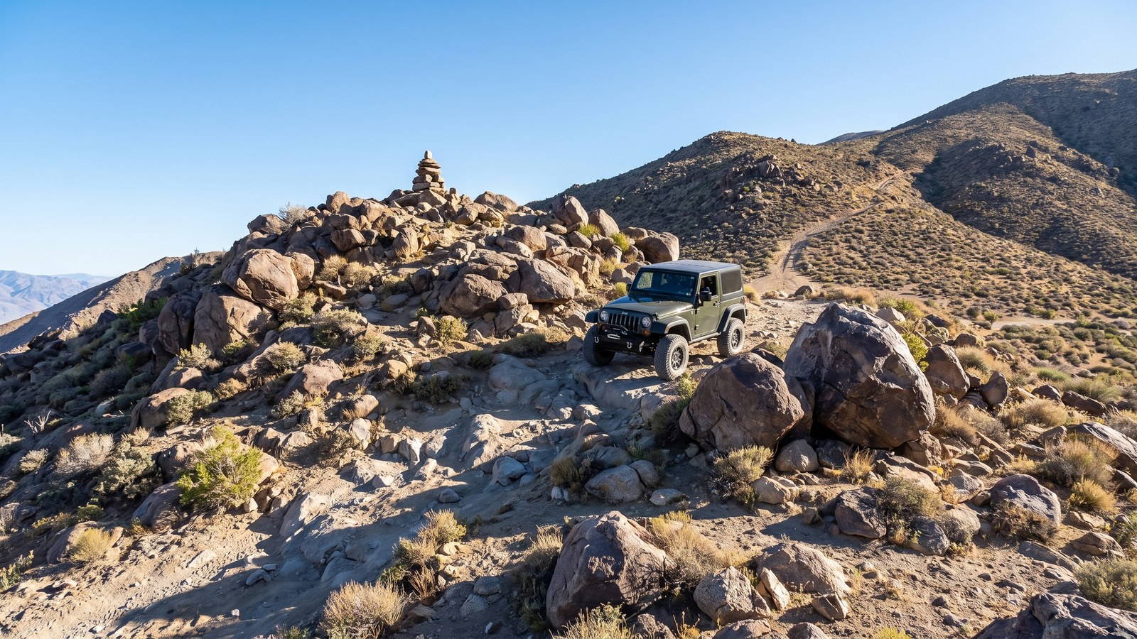 A rocky, boulder-strewn 4x4 pass in the Panamint Range with a stacked-rock grave marker and a rugged desert vehicle cresting the summit under a clear blue sky